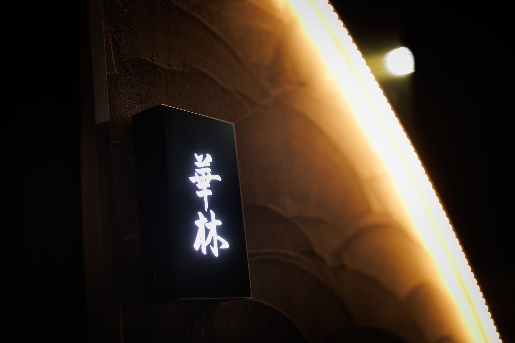 夜の外壁に取り付けられた縦長の看板が白い光で輝き、暖かな照明に照らされた石風の壁が暗闇に浮かぶ外観