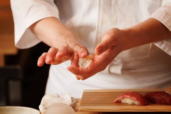 白い調理服の料理人の両手で握り寿司を握る工程の接写。手前の木台には出来上がったマグロの握りが置かれる