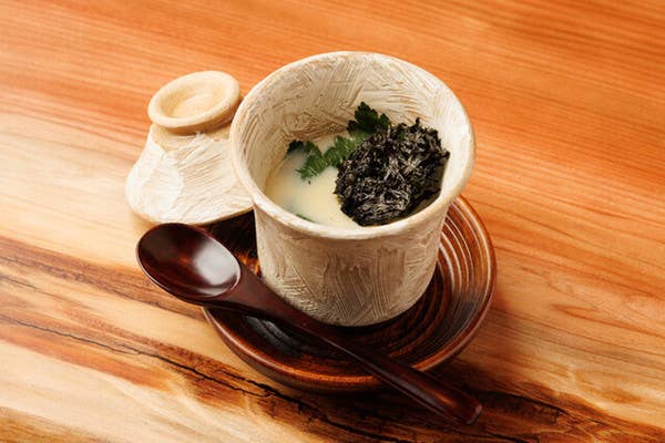 白い刷毛目の器に盛られた茶碗蒸し。木の芽が添えられ、朱色の受皿と木スプーンが置かれる